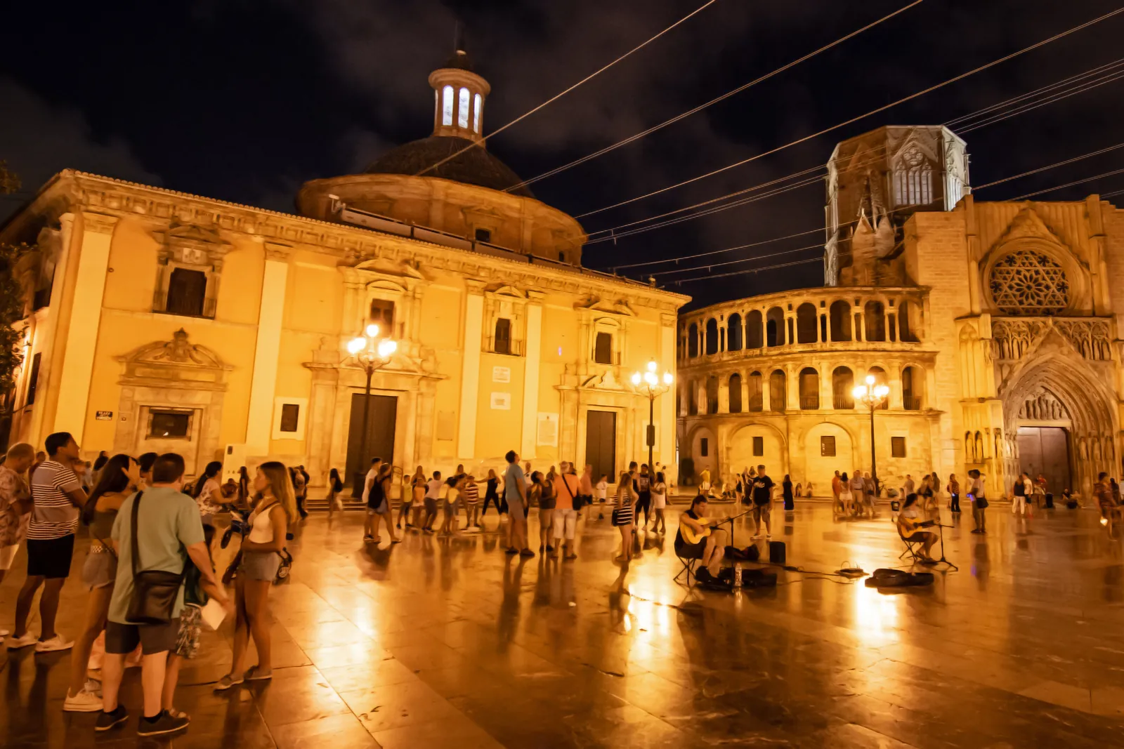 Plaza de la Virgen in Valencia at night, with a crowd on the lamplit square in front of the domed basilica and the cathedral's Gothic doorway